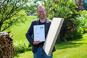 Dieses Bild zeigt Herrn Robert Schwemmer mit der Urkunde des Green Product Award in der rechten hand und einer Schallschutzplatte aus Hanf in der Linken Hand im Hintergrund findet man einen grünen Garten mit einem Holzstoß