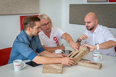 Bild von Geschäftsführer, Werksleiter und Kaufm. Leiter bei der Diskussion über Bauprodukte. Sie sitzen an einem Tisch und berühren die Platten. weiters befinden sich noch 3 Strabag Häferl am Tisch 